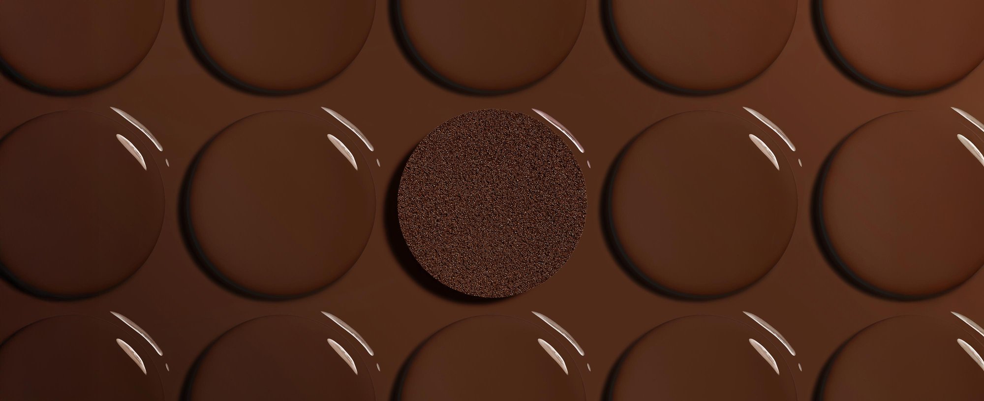 Artistic close-up of smooth, glossy drops of deep brown liquid foundation arranged in rows. In the center, a textured circular foundation cushion is saturated with a matching deep shade, highlighting foundation for deep skin tones.