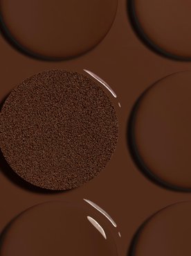 Artistic close-up of smooth, glossy drops of deep brown liquid foundation arranged in rows. In the center, a textured circular foundation cushion is saturated with a matching deep shade, highlighting foundation for deep skin tones.