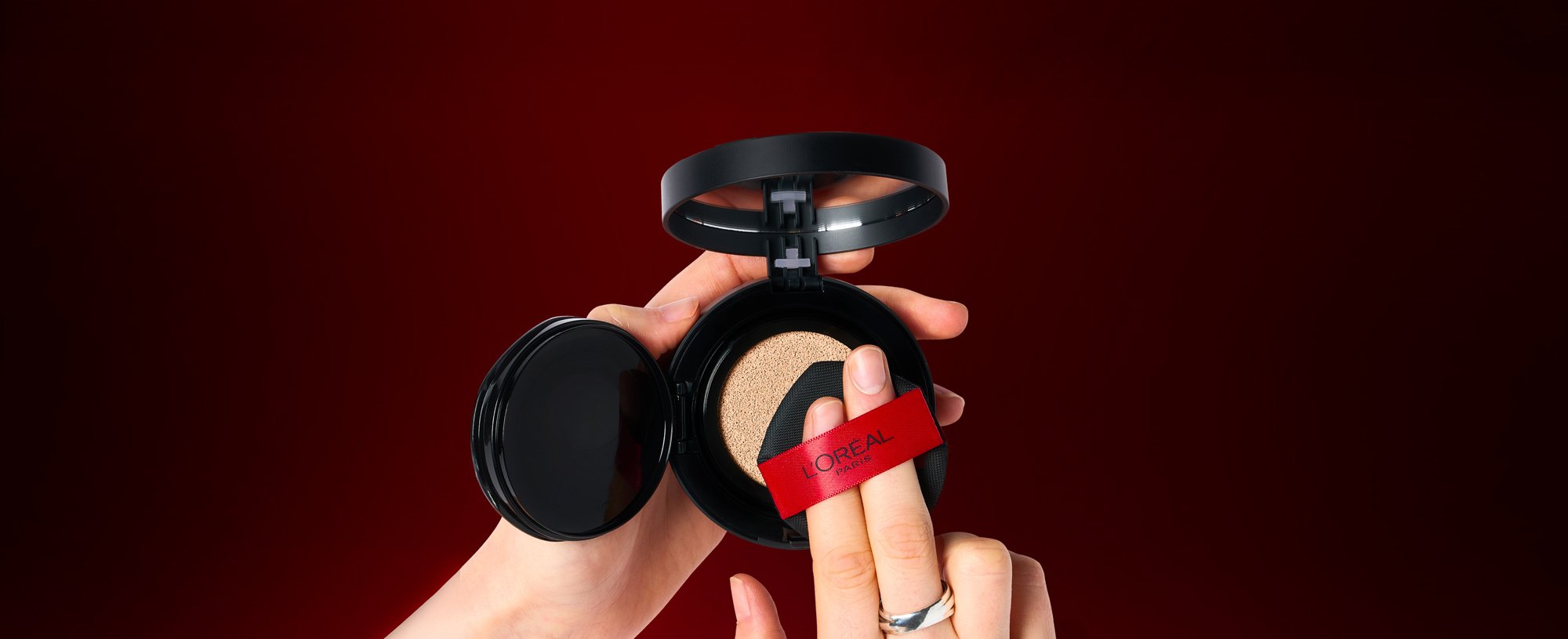 : Gros plan des mains d'une personne tenant un poudrier ouvert de fond de teint cushion L'Oréal Paris. Elle presse un applicateur éponge noir doté d'un ruban rouge L'Oréal dans le coussin imprégné de fond de teint sur un fond rouge foncé.