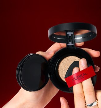 : Gros plan des mains d'une personne tenant un poudrier ouvert de fond de teint cushion L'Oréal Paris. Elle presse un applicateur éponge noir doté d'un ruban rouge L'Oréal dans le coussin imprégné de fond de teint sur un fond rouge foncé.