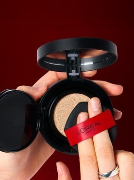 Close-up of a person's hands holding an open L'Oréal Paris cushion foundation compact. They are pressing a black sponge applicator with a red L'Oréal ribbon into the foundation-soaked cushion against a dark red background.