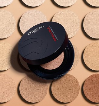 Un poudrier noir élégant de fond de teint cushion Infaillible de L'Oréal Paris présenté ouvert, révélant son miroir intérieur et une houppette applicatrice noire dotée d'un ruban rouge emblématique. Le produit est exposé sur un fond dégradé rouge foncé.
