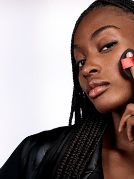 : Une femme noire aux longues tresses foncées et à la peau éclatante applique du maquillage sur sa joue à l'aide d'une éponge applicatrice noire L'Oréal Paris. Elle porte une veste en cuir noir et regarde directement l'objectif sur un fond blanc uni.
