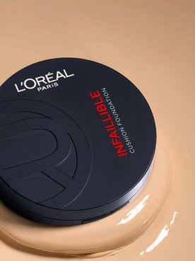 A vertical shot of a closed, matte black L'Oréal Paris Infallible Cushion Foundation compact. The product is resting on a large, smooth puddle of creamy liquid foundation in a light beige shade.