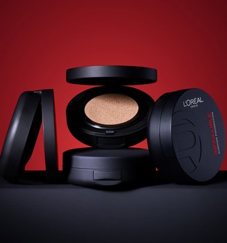 Une collection de quatre poudriers de fond de teint cushion Infaillible de L'Oréal Paris disposés sur une surface noire sur un fond rouge vif. Un poudrier au centre est ouvert, révélant le coussin imprégné de fond de teint, tandis que les autres sont fermés, mettant en valeur l'élégant emballage noir et le nom du produit.