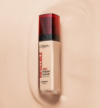 A bottle of L'Oréal Infallible 32H Fresh Wear foundation with a red cap, lying on a smooth, liquid foundation smear of a matching beige shade.