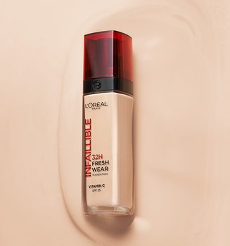 Un flacon de fond de teint Infallible 32H Fresh Wear de L'Oréal avec un bouchon rouge, posé sur une trace de fond de teint liquide d'une teinte beige assortie.