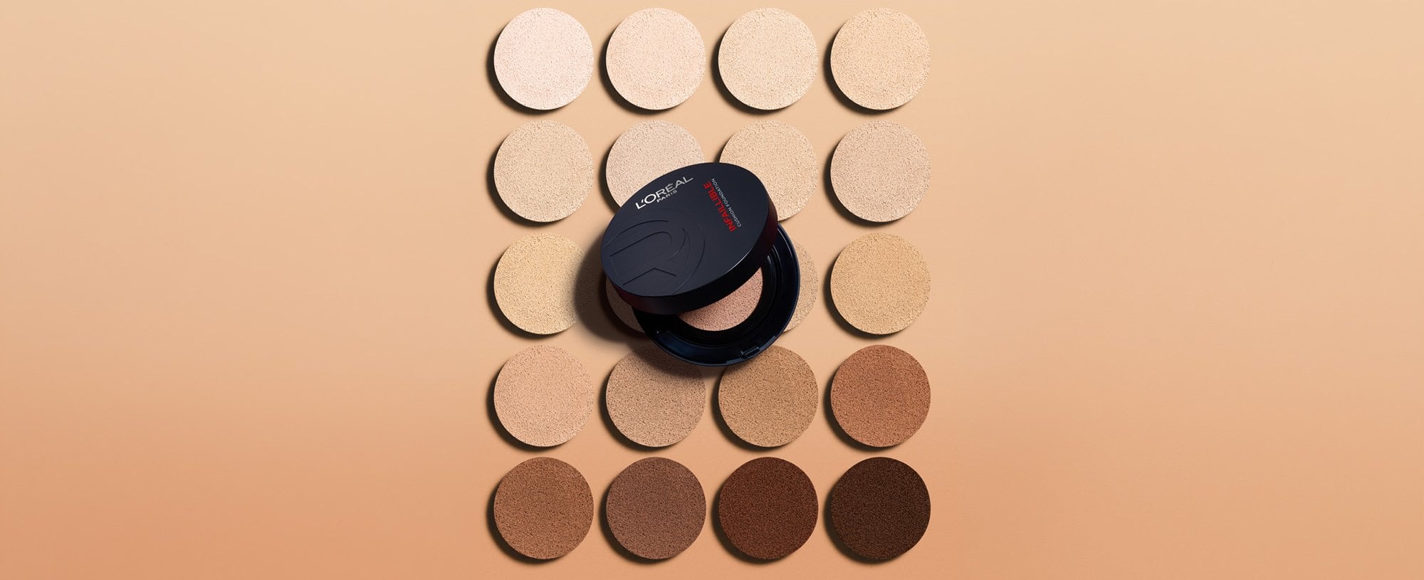 A circular black L'Oréal compact placed in the center of a grid of foundation shade swatches, ranging from very fair to deep brown.