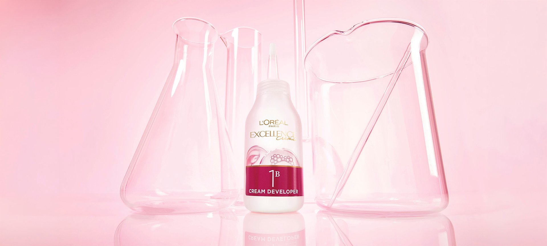 A bottle of L'Oréal Paris Excellence Crème 1B Color Revealing Cream sitting on a pink reflective surface among glass laboratory beakers.