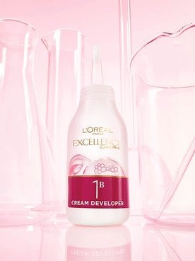 Un flacon de L'Oréal Paris Excellence Crème 1B Révélateur Crème posé sur une surface réfléchissante rose parmi des béchers de laboratoire en verre.