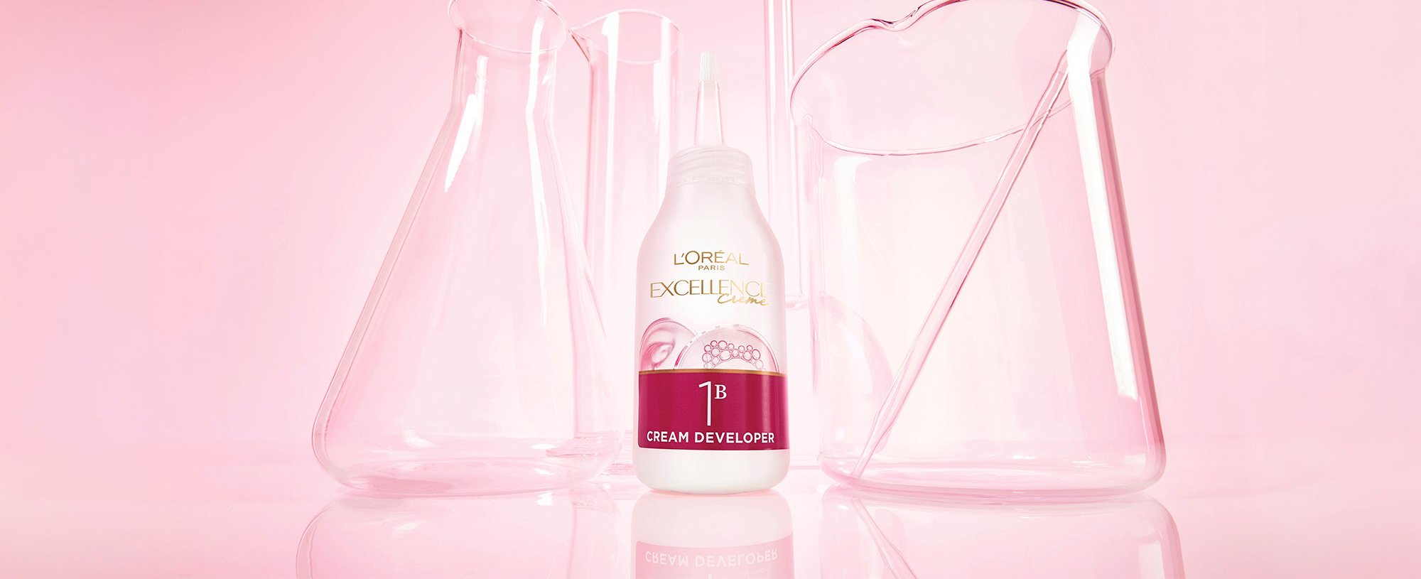 A bottle of L'Oréal Paris Excellence Crème 1B Color Revealing Cream sitting on a pink reflective surface among glass laboratory beakers.