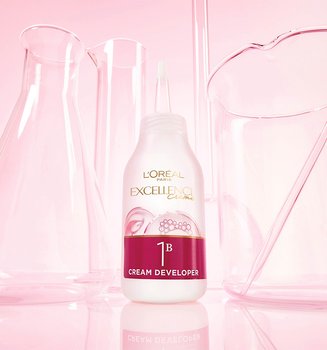A bottle of L'Oréal Paris Excellence Crème 1B Color Revealing Cream sitting on a pink reflective surface among glass laboratory beakers.