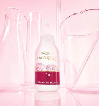 Un flacon de L'Oréal Paris Excellence Crème 1B Révélateur Crème posé sur une surface réfléchissante rose parmi des béchers de laboratoire en verre.
