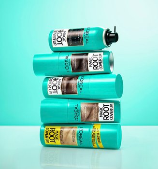 A stack of five L'Oréal Magic Root Cover Up spray cans in shades from black to blonde, set against a vibrant turquoise background.