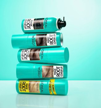Une pile de cinq sprays L'Oréal Magic Root Cover Up dans des teintes allant du noir au blond, sur un fond turquoise éclatant.