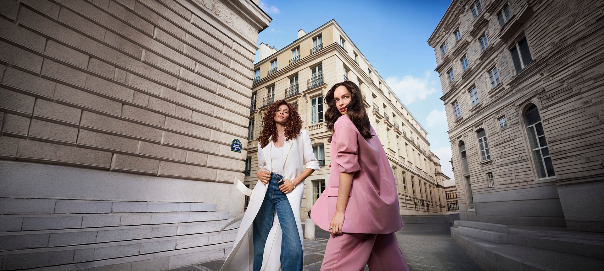Deux femmes élégantes, l'une en long manteau blanc et l'autre en tailleur rose, posant dans une rue historique aux architectures classiques.