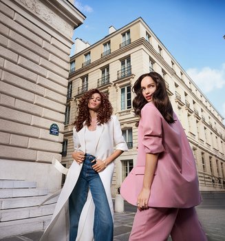 Two stylish women, one in a long white coat and one in a pink suit, posing on a historic city street with classic architecture.