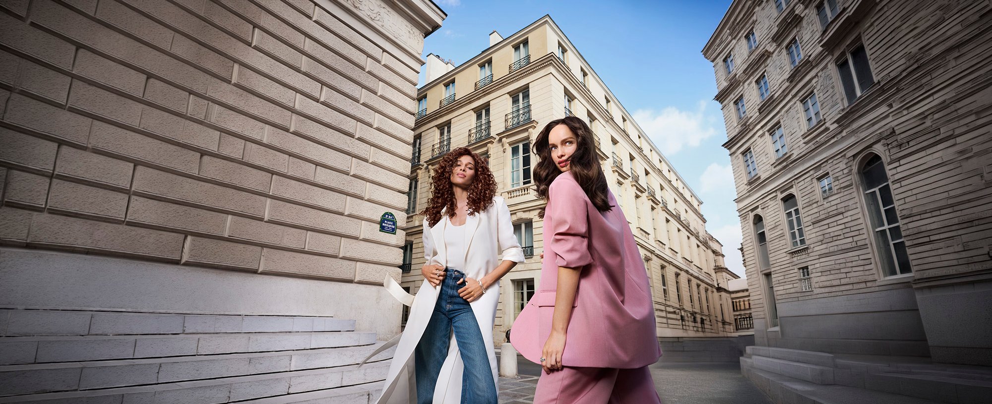 Two stylish women, one in a long white coat and one in a pink suit, posing on a historic city street with classic architecture.