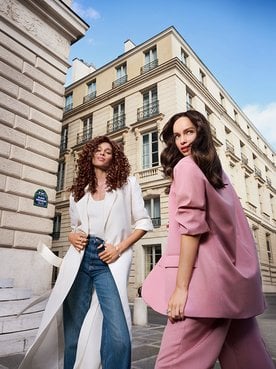 Two stylish women, one in a long white coat and one in a pink suit, posing on a historic city street with classic architecture.
