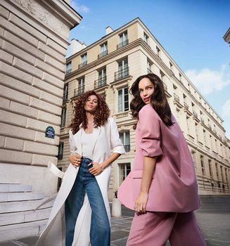 Two stylish women, one in a long white coat and one in a pink suit, posing on a historic city street with classic architecture.