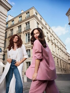 Deux femmes élégantes, l'une en long manteau blanc et l'autre en tailleur rose, posant dans une rue historique aux architectures classiques.