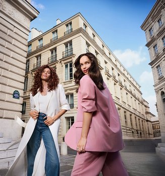 Deux femmes élégantes, l'une en long manteau blanc et l'autre en tailleur rose, posant dans une rue historique aux architectures classiques.