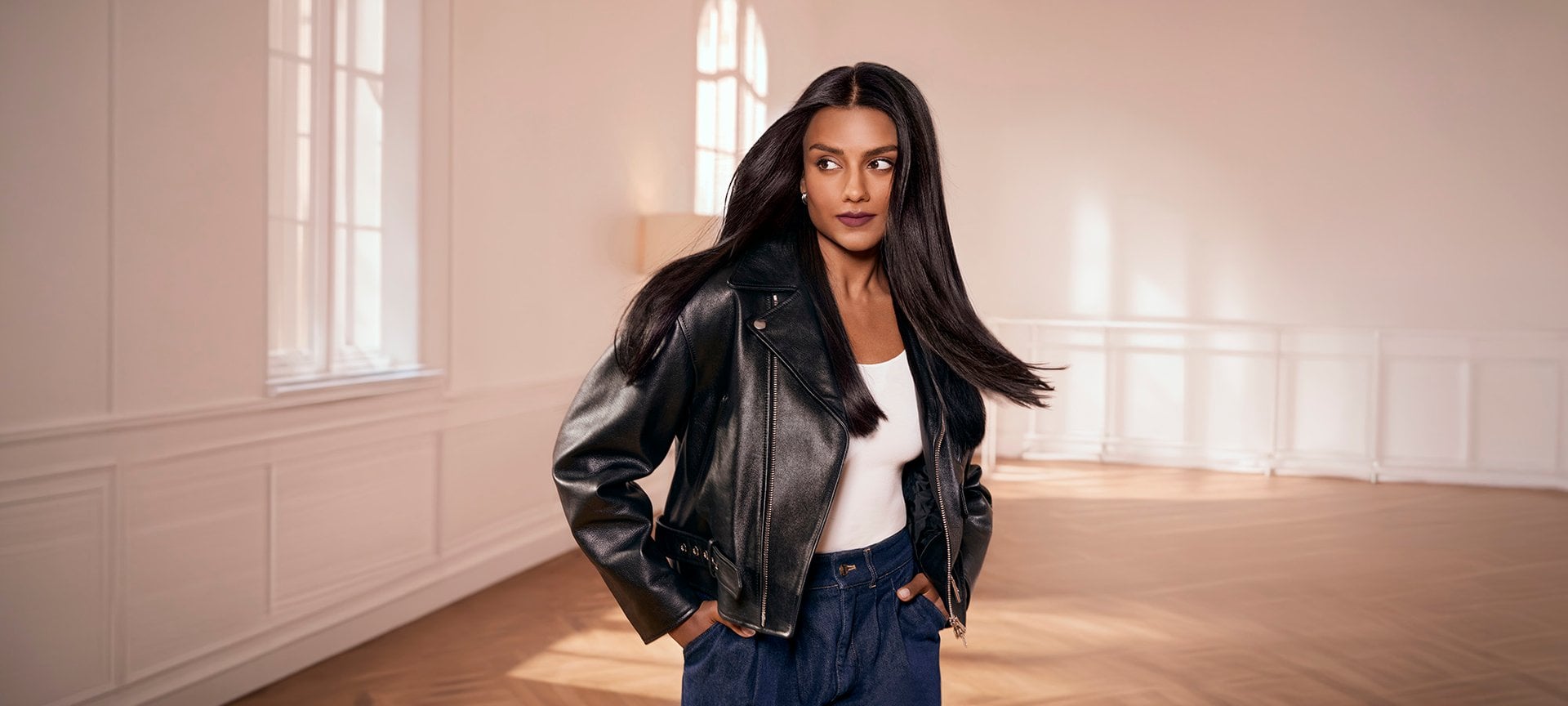 Simone Ashley avec de longs cheveux noirs lisses, portant une veste en cuir noir, un haut blanc et un jean, posant dans une pièce lumineuse et spacieuse avec du parquet.