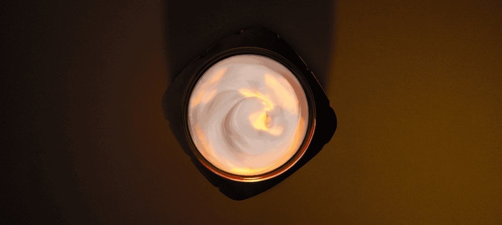 Artistic overhead photography of an open L'Oréal Paris skincare jar. The black, squared-off jar contains a rich white cream with an elegant swirled texture. A warm golden light illuminates the center of the cream from above, creating a glowing effect that highlights the product's luxurious consistency. The composition is set against a dark, minimalist chocolate-brown background.