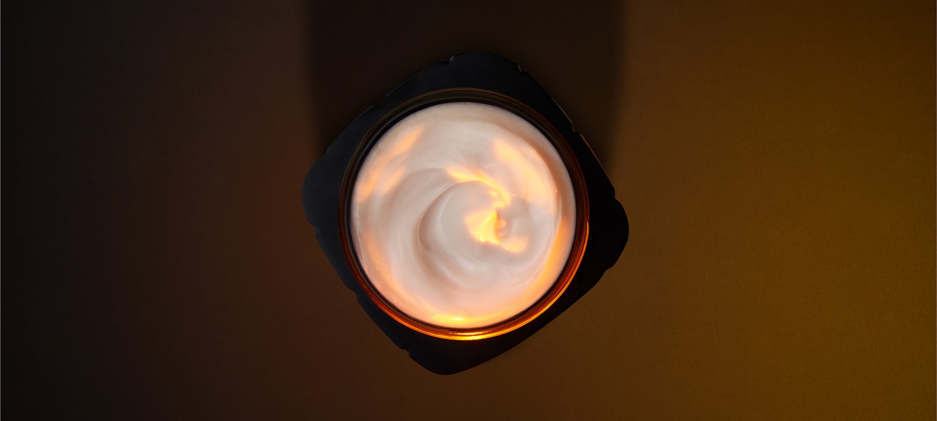 Artistic overhead photography of an open L'Oréal Paris skincare jar. The black, squared-off jar contains a rich white cream with an elegant swirled texture. A warm golden light illuminates the center of the cream from above, creating a glowing effect that highlights the product's luxurious consistency. The composition is set against a dark, minimalist chocolate-brown background.