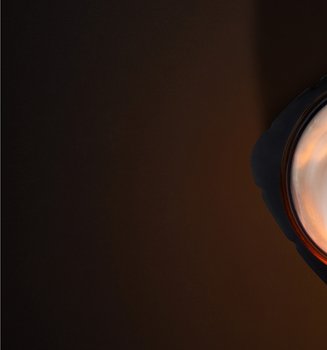 Artistic overhead photography of an open L'Oréal Paris skincare jar. The black, squared-off jar contains a rich white cream with an elegant swirled texture. A warm golden light illuminates the center of the cream from above, creating a glowing effect that highlights the product's luxurious consistency. The composition is set against a dark, minimalist chocolate-brown background.