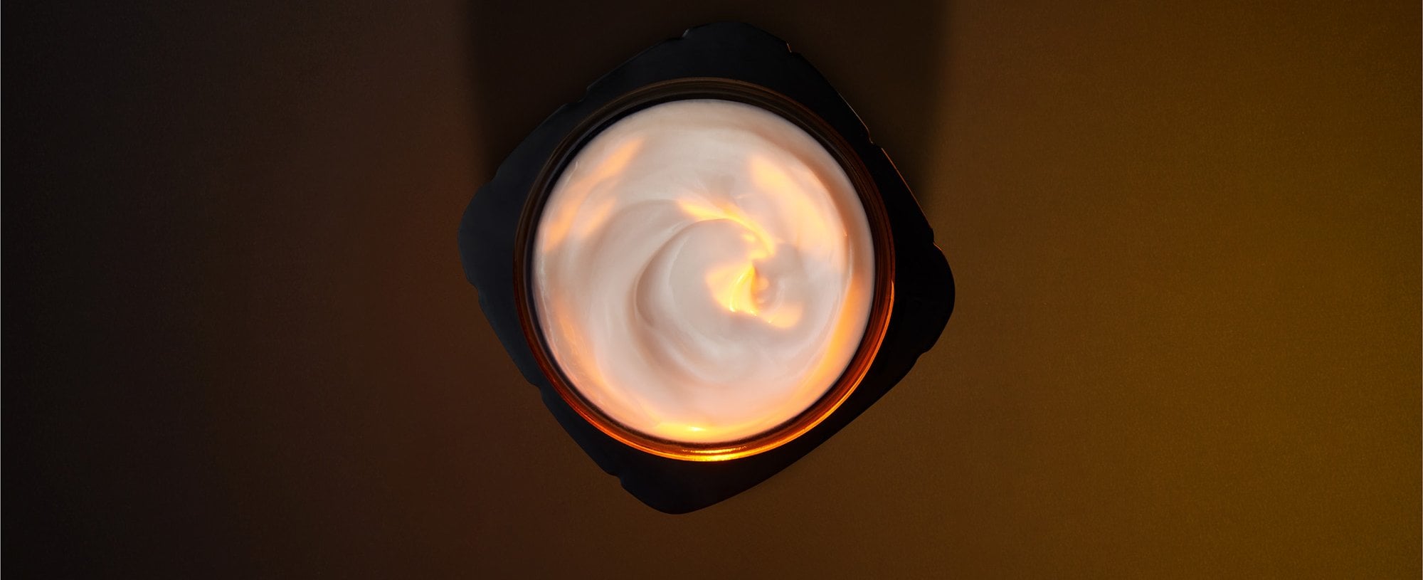 Photographie artistique en vue de dessus d'un pot de soin L'Oréal Paris ouvert. Le pot noir, aux contours anguleux, contient une crème blanche onctueuse présentant un élégant mouvement de tourbillon. Une source de lumière chaude et dorée illumine directement le cœur de la texture, créant un effet de lueur interne et soulignant la richesse du produit. L'ensemble est mis en scène sur un fond sombre et minimaliste aux tons brun chocolat.