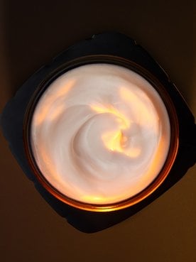 Artistic overhead photography of an open L'Oréal Paris skincare jar. The black, squared-off jar contains a rich white cream with an elegant swirled texture. A warm golden light illuminates the center of the cream from above, creating a glowing effect that highlights the product's luxurious consistency. The composition is set against a dark, minimalist chocolate-brown background.