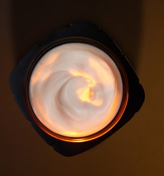 Photographie artistique en vue de dessus d'un pot de soin L'Oréal Paris ouvert. Le pot noir, aux contours anguleux, contient une crème blanche onctueuse présentant un élégant mouvement de tourbillon. Une source de lumière chaude et dorée illumine directement le cœur de la texture, créant un effet de lueur interne et soulignant la richesse du produit. L'ensemble est mis en scène sur un fond sombre et minimaliste aux tons brun chocolat.