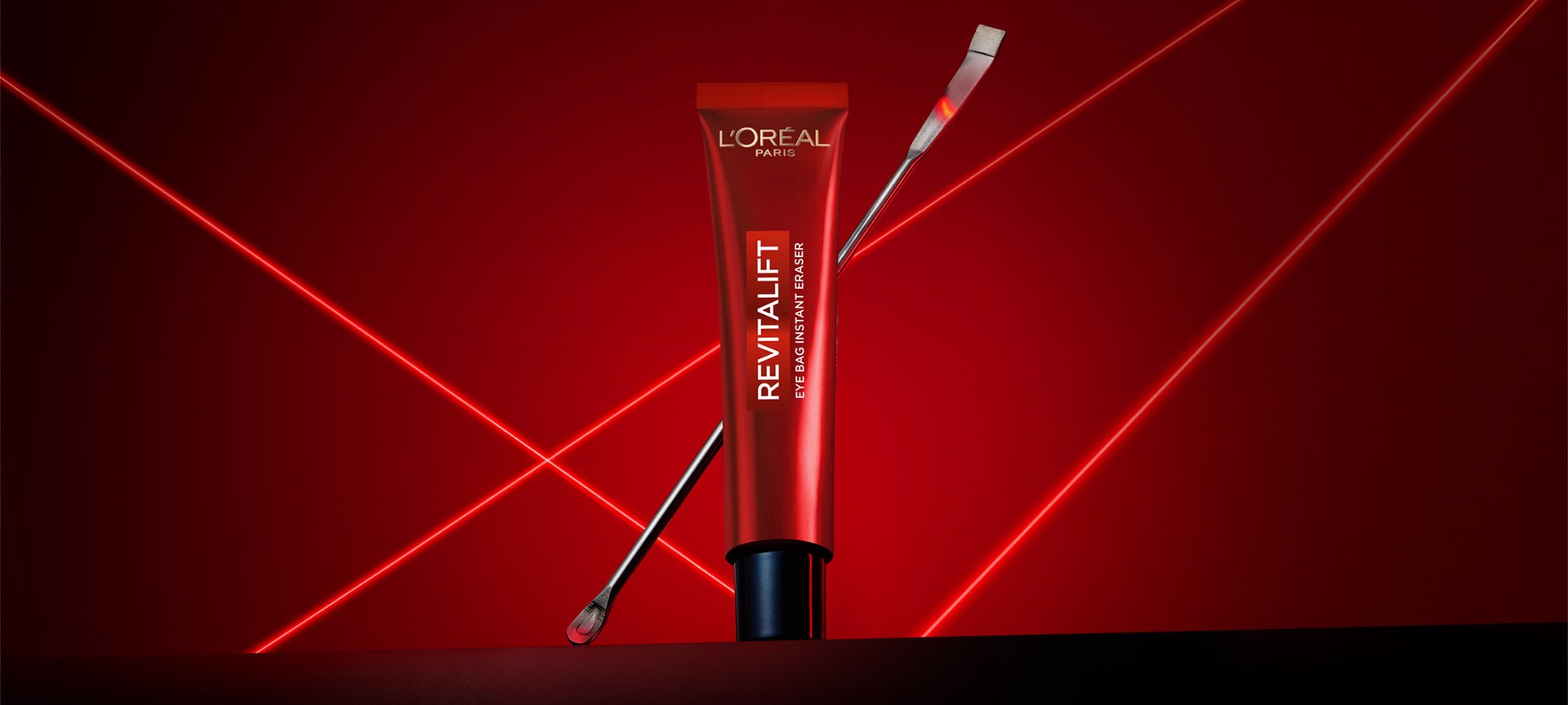 Une photographie publicitaire professionnelle du soin Revitalift Eye Bag Instant Eraser de L'Oréal Paris. Un tube rouge élégant est posé verticalement au centre sur une surface sombre et réfléchissante, avec une fine spatule métallique de laboratoire appuyée en diagonale derrière lui. La mise en scène se détache sur un fond rouge profond dynamisé par des lignes laser vives et lumineuses qui s'entrecroisent en forme de 'X', créant une ambiance technologique, clinique et scientifique qui souligne la précision et l'expertise dermatologique du produit.