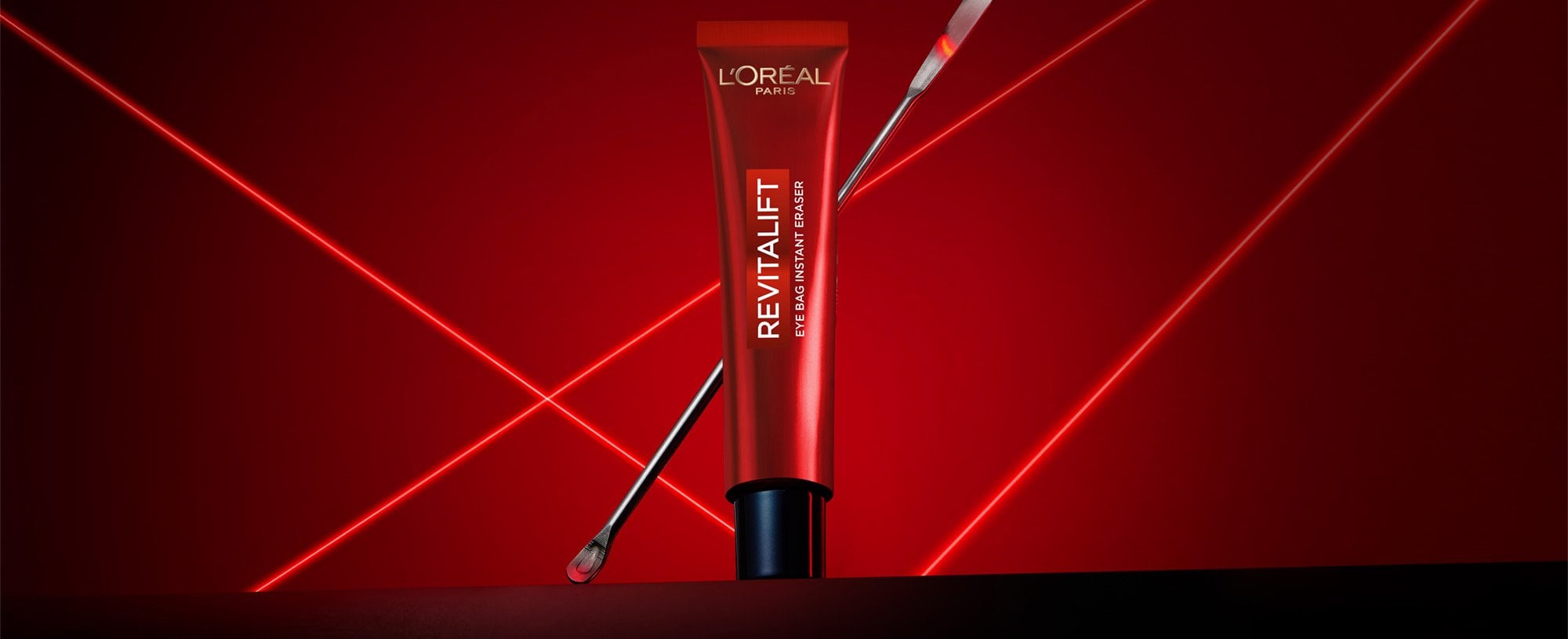 Une photographie publicitaire professionnelle du soin Revitalift Eye Bag Instant Eraser de L'Oréal Paris. Un tube rouge élégant est posé verticalement au centre sur une surface sombre et réfléchissante, avec une fine spatule métallique de laboratoire appuyée en diagonale derrière lui. La mise en scène se détache sur un fond rouge profond dynamisé par des lignes laser vives et lumineuses qui s'entrecroisent en forme de 'X', créant une ambiance technologique, clinique et scientifique qui souligne la précision et l'expertise dermatologique du produit.