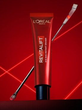 A professional product photography of the L'Oréal Paris Revitalift Eye Bag Instant Eraser. A sleek red tube stands upright in the center on a dark, reflective surface, with a thin metallic laboratory spatula leaning diagonally behind it. The composition is set against a deep red background illuminated by vibrant, glowing red laser lines that crisscross in an 'X' pattern, creating a high-tech, clinical, and scientific atmosphere that emphasizes the precision and dermatological expertise of the skincare product