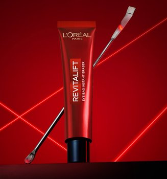 Une photographie publicitaire professionnelle du soin Revitalift Eye Bag Instant Eraser de L'Oréal Paris. Un tube rouge élégant est posé verticalement au centre sur une surface sombre et réfléchissante, avec une fine spatule métallique de laboratoire appuyée en diagonale derrière lui. La mise en scène se détache sur un fond rouge profond dynamisé par des lignes laser vives et lumineuses qui s'entrecroisent en forme de 'X', créant une ambiance technologique, clinique et scientifique qui souligne la précision et l'expertise dermatologique du produit.