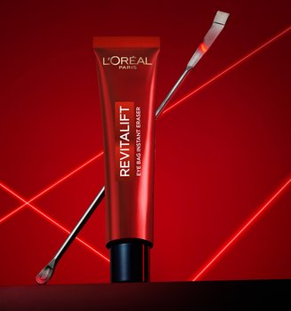 Une photographie publicitaire professionnelle du soin Revitalift Eye Bag Instant Eraser de L'Oréal Paris. Un tube rouge élégant est posé verticalement au centre sur une surface sombre et réfléchissante, avec une fine spatule métallique de laboratoire appuyée en diagonale derrière lui. La mise en scène se détache sur un fond rouge profond dynamisé par des lignes laser vives et lumineuses qui s'entrecroisent en forme de 'X', créant une ambiance technologique, clinique et scientifique qui souligne la précision et l'expertise dermatologique du produit.