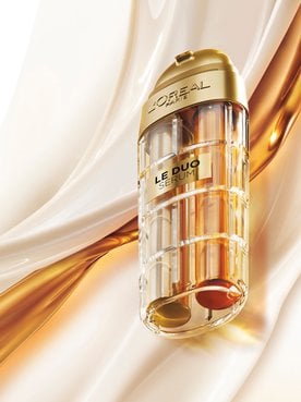 Image publicitaire du produit L'Oréal Paris Le Duo Serum. Le flacon luxueux, aux accents dorés et au corps transparent révélant deux formules distinctes (une claire et une ambrée), est présenté en diagonale. L'arrière-plan se compose de vagues onctueuses et satinées de textures blanches et dorées qui s'entrelacent, symbolisant la puissance et la richesse du mélange de sérums.