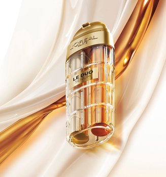 Image publicitaire du produit L'Oréal Paris Le Duo Serum. Le flacon luxueux, aux accents dorés et au corps transparent révélant deux formules distinctes (une claire et une ambrée), est présenté en diagonale. L'arrière-plan se compose de vagues onctueuses et satinées de textures blanches et dorées qui s'entrelacent, symbolisant la puissance et la richesse du mélange de sérums.
