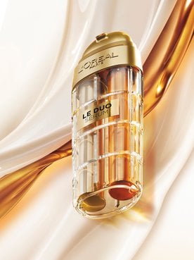 Commercial image of the L'Oréal Paris Le Duo Serum. The luxurious bottle, with gold accents and a transparent body revealing two distinct formulas (one clear and one amber), is presented at a diagonal angle. The background consists of smooth, silky waves of white and golden textures swirling together, symbolizing the power and richness of the serum blend.
