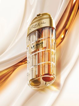 Commercial image of the L'Oréal Paris Le Duo Serum. The luxurious bottle, with gold accents and a transparent body revealing two distinct formulas (one clear and one amber), is presented at a diagonal angle. The background consists of smooth, silky waves of white and golden textures swirling together, symbolizing the power and richness of the serum blend.
