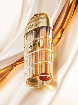 Image publicitaire du produit L'Oréal Paris Le Duo Serum. Le flacon luxueux, aux accents dorés et au corps transparent révélant deux formules distinctes (une claire et une ambrée), est présenté en diagonale. L'arrière-plan se compose de vagues onctueuses et satinées de textures blanches et dorées qui s'entrelacent, symbolisant la puissance et la richesse du mélange de sérums.
