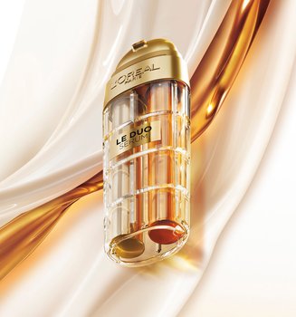 Image publicitaire du produit L'Oréal Paris Le Duo Serum. Le flacon luxueux, aux accents dorés et au corps transparent révélant deux formules distinctes (une claire et une ambrée), est présenté en diagonale. L'arrière-plan se compose de vagues onctueuses et satinées de textures blanches et dorées qui s'entrelacent, symbolisant la puissance et la richesse du mélange de sérums.