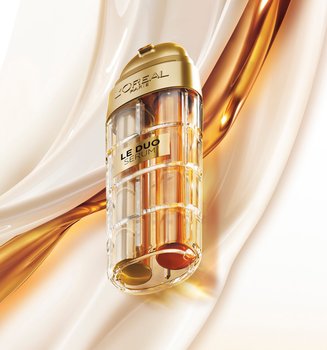 Commercial image of the L'Oréal Paris Le Duo Serum. The luxurious bottle, with gold accents and a transparent body revealing two distinct formulas (one clear and one amber), is presented at a diagonal angle. The background consists of smooth, silky waves of white and golden textures swirling together, symbolizing the power and richness of the serum blend.