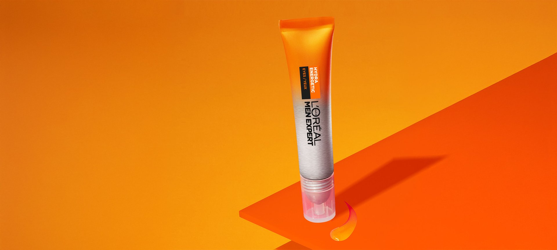 Orange tube of L'Oréal Men Expert Hydrating Cream lying on orange geometric shapes, with a visible product droplet, on an orange background.