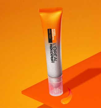 Orange tube of L'Oréal Men Expert Hydrating Cream lying on orange geometric shapes, with a visible product droplet, on an orange background.