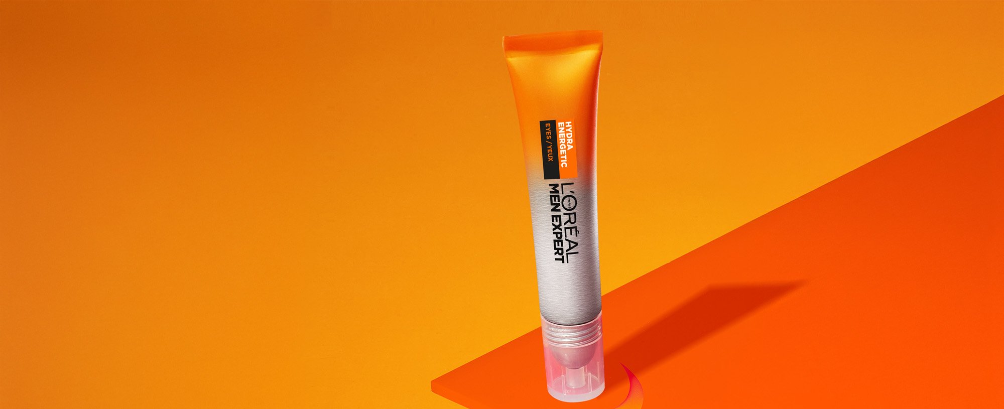 Tube orange de soin L'Oréal Men Expert Hydratation sur des formes géométriques oranges, avec une goutte de produit visible, sur un fond orange