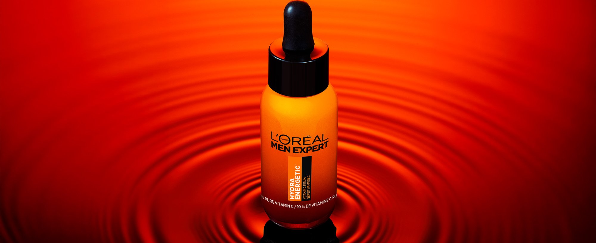 Flacon orange de sérum L'Oréal Men Expert Hydratatic avec pipette noire, sur une surface rouge réfléchissante avec des ondulations.