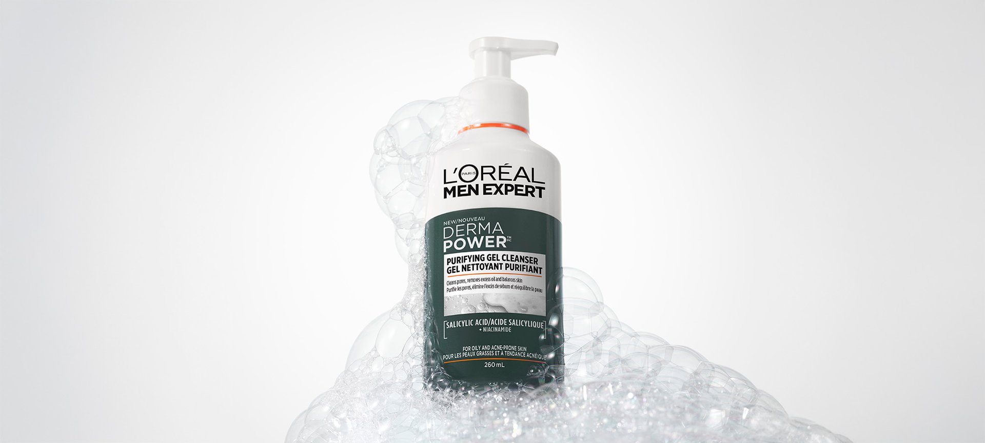 White bottle of L'Oréal Men Expert Derma Power Purifying Gel Cleanser with a pump, surrounded by white soap bubbles on a grey background.